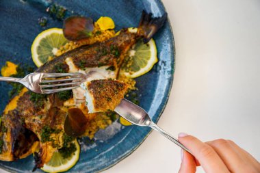 A man holds a fork and a knife with a piece of fried fish, and in the background is a whole fried fish with lemon, sauce and green petals in a plate that stands on the tableA man holds a fork and a knife with a piece of fried fish, and in the backgro