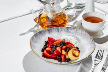 Breakfast. Morning dish of sweet cottage cheese pancakes, ricotta cheese, blueberries and strawberries. Cheesecakes lie in a round, ceramic, light handmade plate. The plate is on a table with a white tablecloth, next to it is a transparent teapot wit