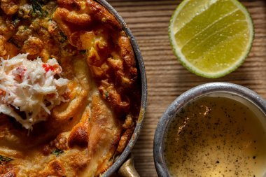 Closed pie with salmon, spinach and cheese, with crabmeat on top. Pie in a metal pan with gilded handles. The saucepan stands on a wooden board, next to it lies half a lime and there is a metal gravy boat with cheese sauce. A board with food stands o