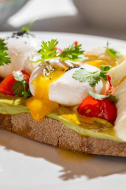 Italian dish. Buckwheat bread bruschetta with mashed avocado, cherry tomatoes, artichokes, parsley leaves and two poached eggs topped with ground truffle. Bruschetta stands on a round, white ceramic plate. Nearby is a champagne glass with a long stem