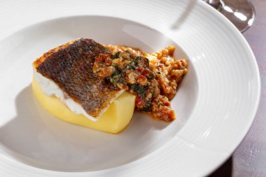 Fried piece of fish steak with mashed potatoes, zucchini, tomatoes, peppers and seaweed in a plate on a wooden table with a glass of wine