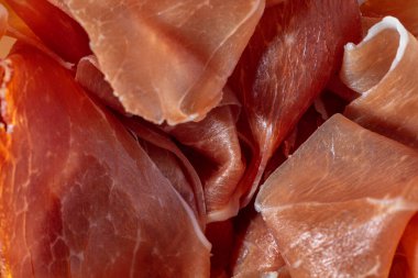 İtalyan jambonlu domuz eti. Porsiyonlar halinde dilimlenmiş ve beyaz, yuvarlak, seramik bir tabağa serpiştirilmiş. Plakası gri, taş bir arka planda duruyor..