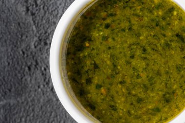 Pesto sosu, beyaz, yuvarlak, seramik bir kasede. Kase gri, taştan bir arka planda duruyor. Yakınlarda yeşil bir fesleğen yaprağı yatıyor..