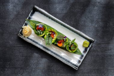 Spring rolls with avocado, cucumber, lettuce, carrots, edible violet petals and wrapped in rice paper. The rolls stand on a bamboo leaf, next to it is pickled ginger and a wasabi cube. Springrolls lie on a rectangular, light, ceramic plate with dark 