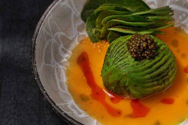 Scallop sashimi under a pillow of avocado slices, topped with a handful of black caviar. Sashimi in yellow tomato sauce with paprika, lettuce sprinkled with turmeric. The dish is in a round, ceramic, brown bowl with a pattern. The plate stands on a g
