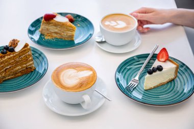 Yaban mersinli cheesecake, çilek ve Napolyon ile aynı masada duran bir çatal ve ikinci tabakta çilek, üçüncü tabakta yabanmersinli ballı kek ve köpüklü iki fincan kapuçino.