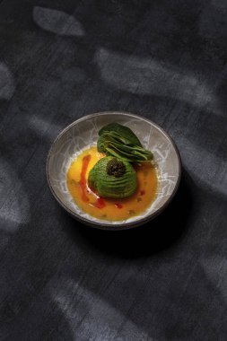 Scallop sashimi under a pillow of avocado slices, topped with a handful of black caviar. Sashimi in yellow tomato sauce with paprika, lettuce sprinkled with turmeric. The dish is in a round, ceramic, brown bowl with a pattern. The plate stands on a g
