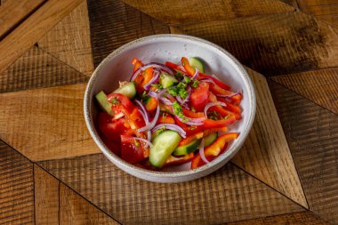 Spring salad with fresh vegetables. Salad with fresh tomatoes, paprika, cucumber, red onion, chopped cilantro and olive oil. The salad lies in a round, deep, ceramic bowl. The dishes are on a wooden table with a pattern.