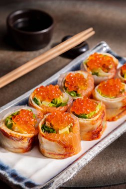 Roll with eel, salmon, red salmon caviar, avocado and salad on a marble plate on the table with bamboo chopsticks and sauce in a saucepan