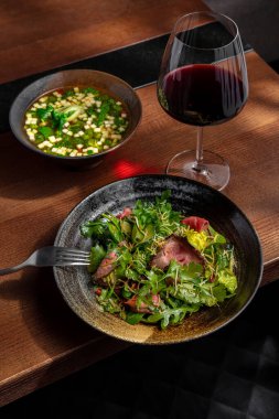 Salad with meat, arugula, frisse salad, cucumber, pepper and slices of sweet potatoes in a plate on the table with a fork, another plate in which is soup, broccoli, basil and cucumber and a glass of red wine
