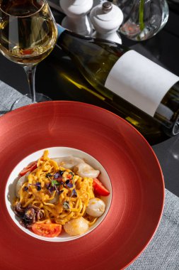 Spaghetti with squid, octopus, sprouts, tomatoes, cheese and sauce in a plate on the table with a glass of wine, a bottle of wine, pepper and salt