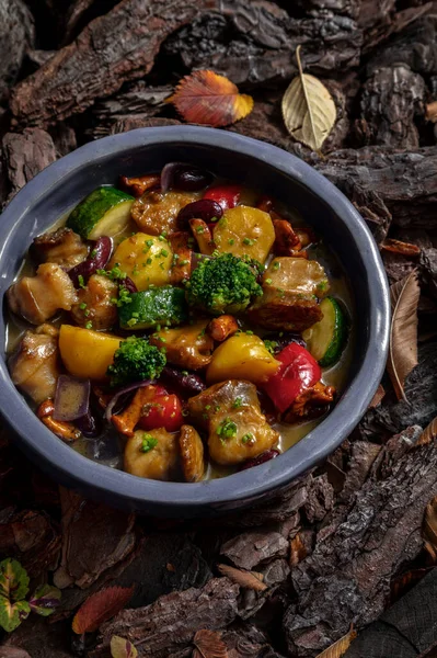 Vegetable stew with chanterelle mushrooms, broccoli, paprika, potatoes, red beans, zucchini and rapanas in a creamy white wine sauce. The stew lies in a blue, round, ceramic plate. The plate stands on a background of tree bark.