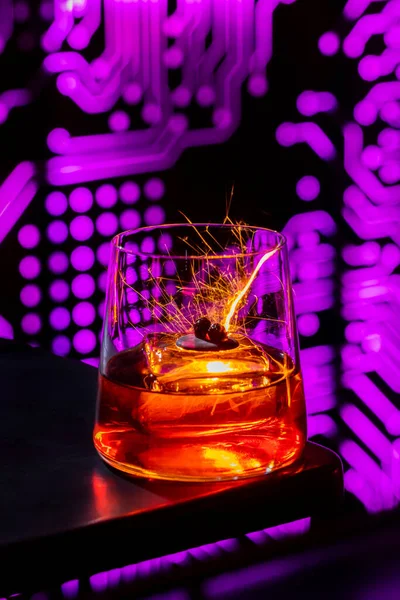 Bourbon whiskey with ice. In transparent. a low glass is poured with bourbon and there is an ice cube. The glass is on the edge of a black table. Behind the glass you can see a luminous pattern in the form of microcircuits. Bengal fire drawing, long