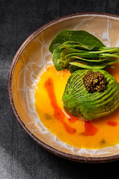 Scallop sashimi under a pillow of avocado slices, topped with a handful of black caviar. Sashimi in yellow tomato sauce with paprika, lettuce sprinkled with turmeric. The dish is in a round, ceramic, brown bowl with a pattern. The plate stands on a g
