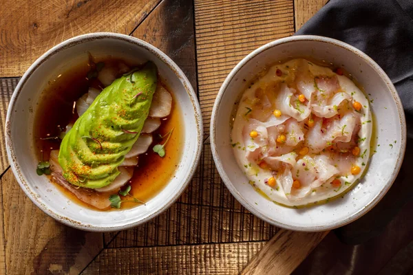 Two course lunch. Sea bass fillet sashimi with avocado and teriyaki sauce and sea bass fillet sashimi with sea buckthorn berries and cream sauce. The food lies in light, ceramic plates. Two plates stand on a wooden table, under one of the plates lies