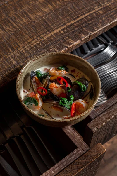 Tom yam soup with coconut milk, mussels, shrimp, chuka seaweed and chili peppers. Soup in a deep, ceramic bowl. The bowl stands on a wooden background, next to a large, old ship's anchor.