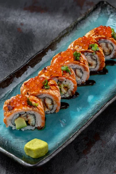 Sushi wrapped in salmon fillet, with red caviar on top and finely chopped green onions, stuffed with avocado and eel in unagi sauce. Sushi stands on a blue, rectangular, ceramic plate with dark sides. Unagi sauce is poured on a plate, there is a squa