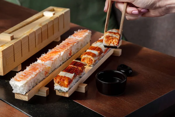 Preparation for rolls with rice, shrimps, eel, yuzo tobiko, tobiko caviar and sauce which is in the machine for making rolls on the table with sauce in a sauce bowl with a man who takes the role of sticks for food