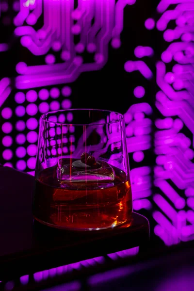 Bourbon whiskey with ice. In transparent. a low glass is poured with bourbon and there is an ice cube. The glass is on the edge of a black table. Behind the glass you can see a luminous pattern in the form of microcircuits.