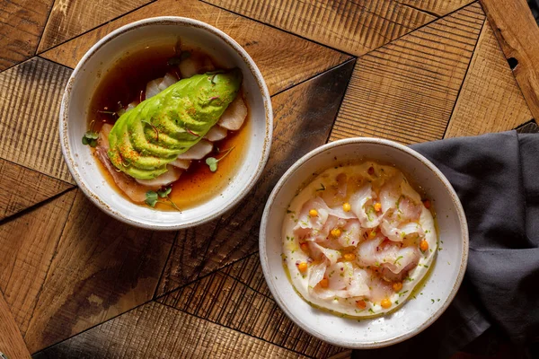 Two course lunch. Sea bass fillet sashimi with avocado and teriyaki sauce and sea bass fillet sashimi with sea buckthorn berries and cream sauce. The food lies in light, ceramic plates. Two plates stand on a wooden table, under one of the plates lies