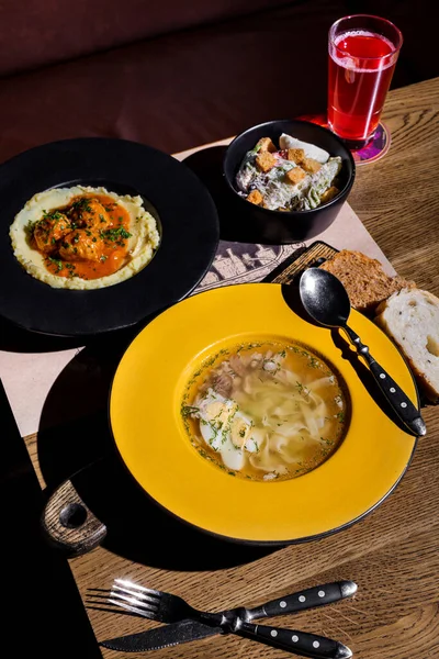 Soup with spaghetti, meat and boiled egg in the first plate with a spoon. Mashed potatoes, meat balls and parsley on the second plate. A bowl in which a boiled egg, crackers, cheese and salad. All this is on the table with cutlery, bread, and juice i