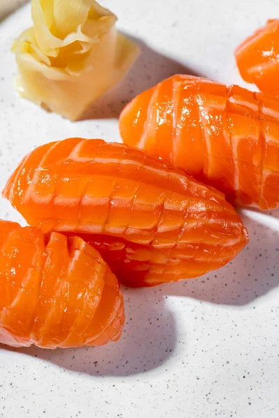 Nigiri with salmon. Large pieces of salmon sashimi lie on top of the rice. Nearby is pickled ginger and a cube of wasabi. The food lies on a round, light, ceramic plate with low sides. The plate stands on a black and beige paper background.