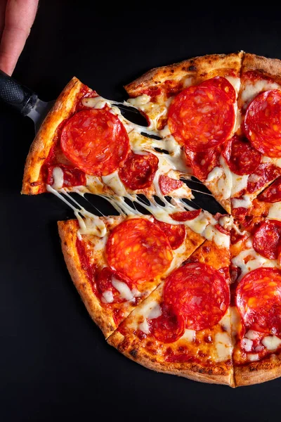 Pizza with pepperoni, sausages, cheese and ketchup on a black background. A man holding a slice of pizza on a spatula