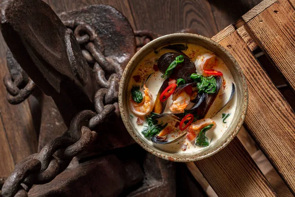 Tom yam soup with coconut milk, mussels, shrimp, chuka seaweed and chili peppers. Soup in a deep, ceramic bowl. The bowl stands on a wooden background, next to a large, old ship\'s anchor.