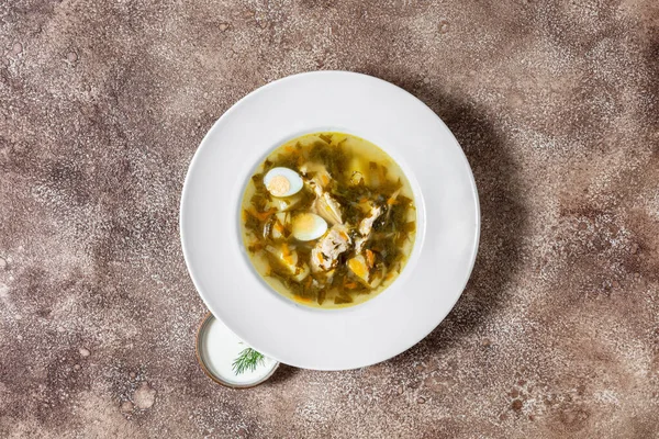 Green borscht soup with sorrel, boiled egg, boiled pork, carrots and potatoes, in a white, round, deep, ceramic plate. The plate stands on a dark, coffee background. Nearby is a small, white, ceramic bowl with sour cream, a sprig of parsley lies on t