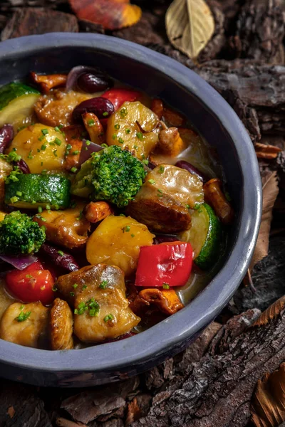 Vegetable stew with chanterelle mushrooms, broccoli, paprika, potatoes, red beans, zucchini and rapanas in a creamy white wine sauce. The stew lies in a blue, round, ceramic plate. The plate stands on a background of tree bark.