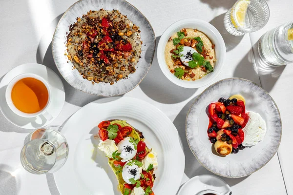 Italian breakfast. There are four morning meals on a table with a white tablecloth. Cheesecakes with berries, oatmeal with berries, oatmeal with nuts and poached egg, bruschetta with avocado, tomato and poached egg. All dishes are in bright ceramic p