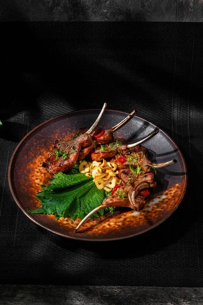 Rack of lamb in sweet chili sauce. grilled. The ribs lie on a lettuce leaf with garlic chips. Sprinkle microgreen sprouts, chili rings and finely chopped parey onion on top. The food lies in a round, brown ceramic plate. The plate stands on a black w