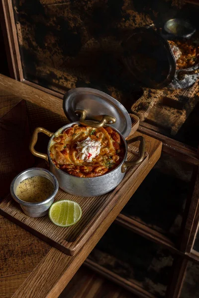 Closed pie with salmon, spinach and cheese, with crabmeat on top. Pie in a metal pan with gilded handles. The saucepan stands on a wooden board, next to it lies half a lime and there is a metal gravy boat with cheese sauce. A board with food stands o