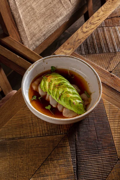 Sea bass fillet sashimi with portion-cut avocado in teriyaki sauce and microgreen sprouts. The food lies in a round, light, ceramic plate with high sides. The plate stands on a wooden table with wood texture patterns.