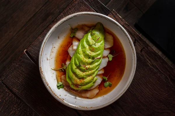 Sea bass fillet sashimi with portion-cut avocado in teriyaki sauce and microgreen sprouts. The food lies in a round, light, ceramic plate with high sides. The plate stands on a wooden table with wood texture patterns.