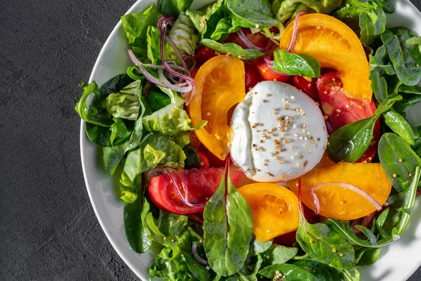 Salad of red and yellow tomatoes, red onion, assorted lettuce, olive oil and Buffalo mozzarella. The salad lies in a white, round, deep, ceramic plate. The plate stands on a gray, stone background.