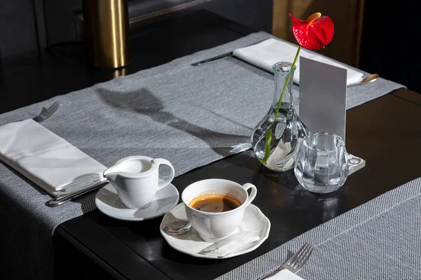 Strong coffee in a cup that stands on a plate on the table with cream in a saucepan, a spoon, a bag of sugar, napkins, cutlery and a vase with a flower