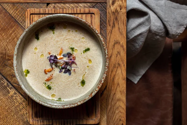 Mushroom creamy mushroom soup with chanterelle mushrooms, pesto sauce and microgreen sprouts. Soup in a deep ceramic bowl. The plate stands on a wooden stand made of light wood, next to it lies a gray, linen fabric. Dishes are on a light, wooden tabl