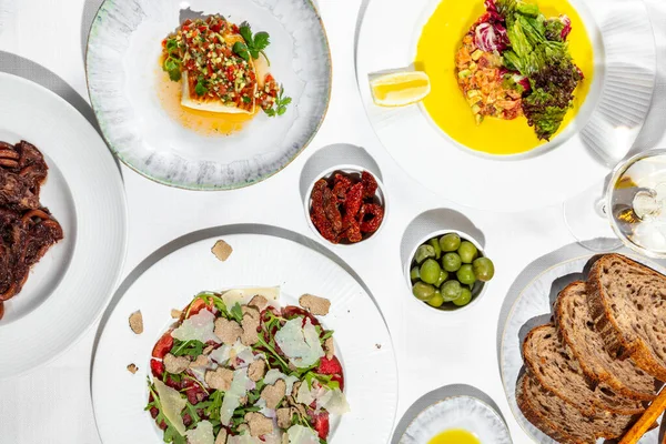 Lunch of Italian cuisine. There are four plates of dishes on a table with a white tablecloth. Bucatini pasta with duck fillet in plum sauce, salmon tartare, perch fillet with tabouleh and veal carpaccio with truffle. The food lies in light, round cer