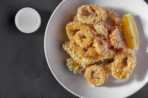 Şarap için aperatif. Soğan halkaları, kırmızı biber, ekmek kırıntılarıyla kızartılmış kabak. Yemek yuvarlak, beyaz, kalın bir tabakta bir dilim limonla birlikte duruyor. Plakası gri, taş bir zeminde duruyor. Yakınlarda bir kase sarımsaklı yoğurt sosu var..