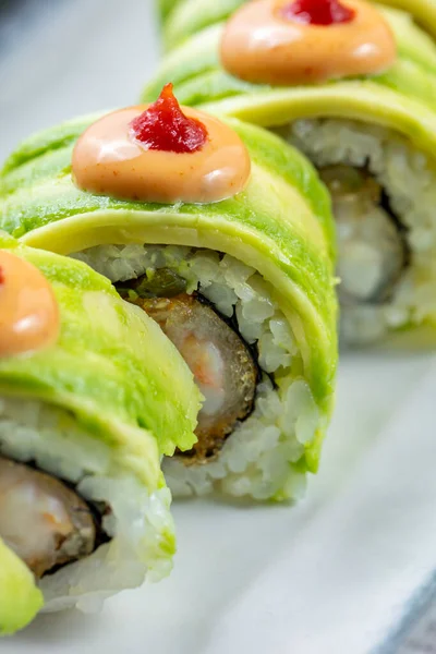 Sushi with rice wrapped in avocado and stuffed with tempura tiger shrimp. On top of the avocado is Japanese chili mayonnaise. Sushi lies on a rectangular, ceramic, light plate. The plate stands on a gray stone background.