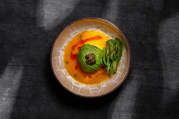 Scallop sashimi under a pillow of avocado slices, topped with a handful of black caviar. Sashimi in yellow tomato sauce with paprika, lettuce sprinkled with turmeric. The dish is in a round, ceramic, brown bowl with a pattern. The plate stands on a g