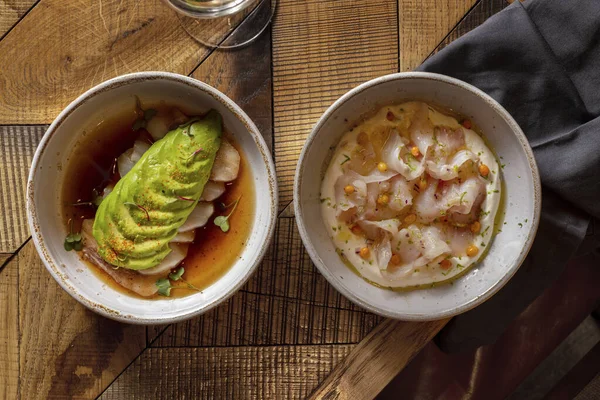 Two course lunch. Sea bass fillet sashimi with avocado and teriyaki sauce and sea bass fillet sashimi with sea buckthorn berries and cream sauce. The food lies in light, ceramic plates. Two plates stand on a wooden table, under one of the plates lies