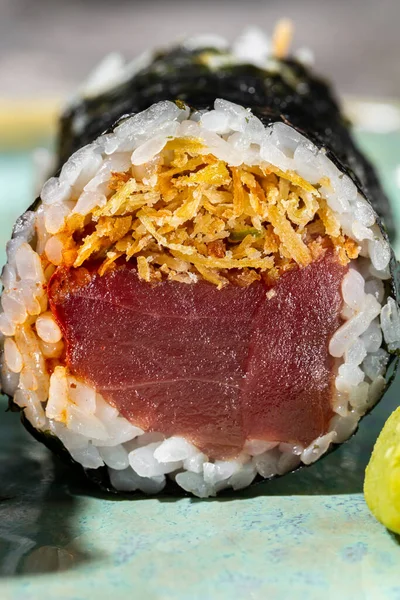 Big roll with tuna. rice, deep-fried onion chips and wrapped in nori seaweed. Two rolls lie on a round, turquoise, ceramic plate with low sides. On the plate, next to the sushi is wasabi and pickled ginger. The plate stands on a gray stone background