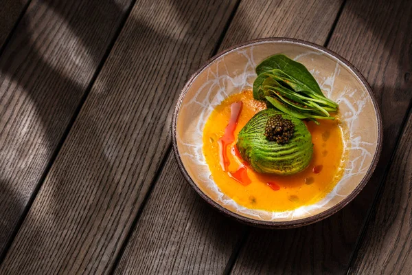 Scallop sashimi under a pillow of avocado slices, topped with a handful of black caviar. Sashimi in yellow tomato sauce with paprika, lettuce sprinkled with turmeric. The dish is in a round, ceramic, brown bowl with a pattern. The plate stands on a b