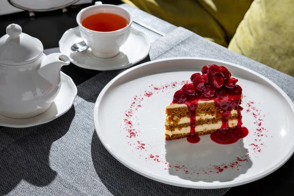 Slice of cake with cherries and cherry jam in a plate on the table with a cup of tea and a teapot