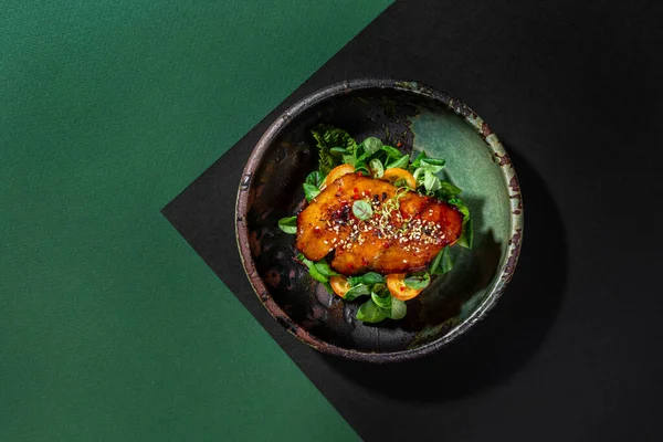 Brioche bun with lettuce, kumquat, microgreen sprouts, on top is sea bass fillet in unagi sauce, sprinkled with sesame seeds and lime zest. The food lies in a deep, ceramic bowl. The bowl stands on paper green and black backgrounds.
