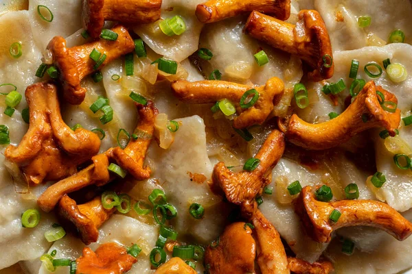 Vareniki with mashed potatoes and chanterelle mushrooms stewed in white wine on top. Finely chopped green onions are sprinkled on top of the dish. Pierogi lie in a round, ceramic plate with high sides. The cook, in a striped apron, holds a plate in h