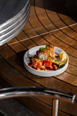 Avokado, tofu ve Philadelphia peyniri ile doldurulmuş, somon balığına sarılı, unagi soslu, ince doğranmış yeşil soğan ve limonlu. Suşi beyaz, yuvarlak, seramik bir tabağın üzerinde duruyor, yanında da turşulu g var.