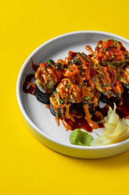 Salmon sushi wrapped in rice and nori seaweed, topped with eel popcorn balls in air dough, unagi sauce and chili sauce, finely chopped green onions are sprinkled on top. Sushi lies on a white, ceramic plate with a pattern. The plate stands on a yello